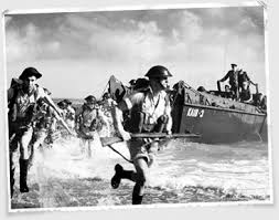 Black and white image of soldiers landing on a beach from a landing craft, carrying rifles and wearing helmets.